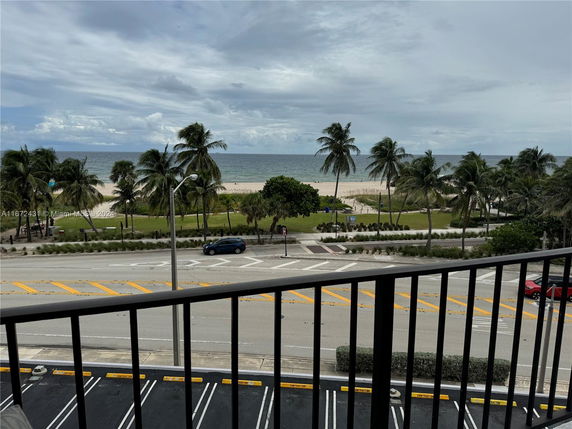 View of beach and ocean from a balcony with palm trees and a road in the foreground.