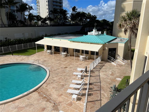 View of a small building with a green-tiled roof near a swimming pool, surrounded by high-rise buildings.