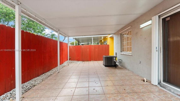 Covered outdoor patio area with tiled flooring and an air conditioning unit, enclosed by a red wooden fence.