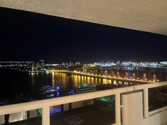 Nighttime cityscape view from a high balcony overlooking a waterfront and illuminated city lights.