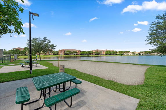Outdoor area with a sand volleyball court by a lake, picnic tables, and nearby residential buildings.