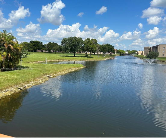 Wide view of a lake with grassy banks and trees, buildings visible in the background.