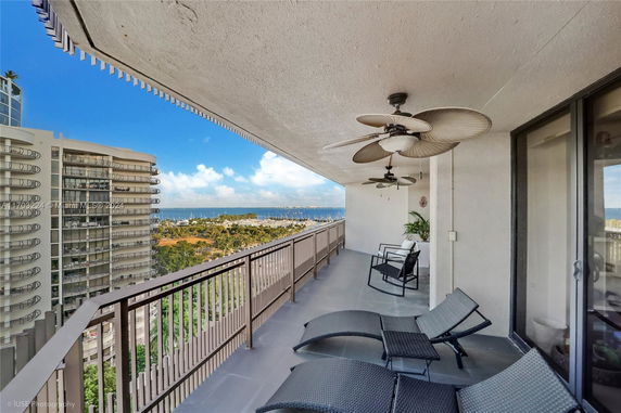 Balcony view with lounge chairs and ceiling fans overlooking buildings and distant ocean.