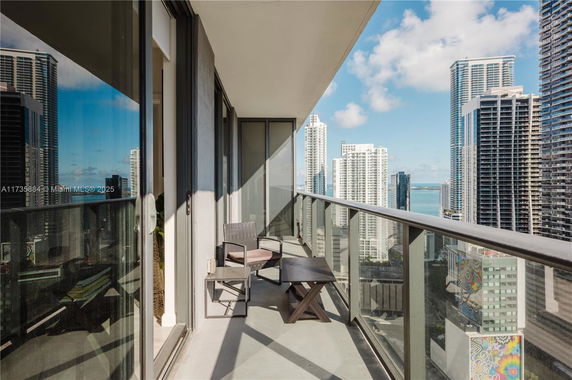 High-rise balcony view with chairs and table overlooking city skyscrapers and water in the distance.