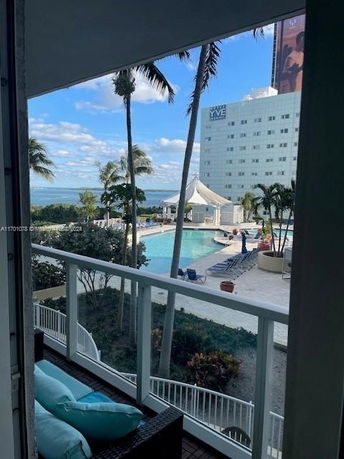 View from a building balcony, overlooking a pool area and the ocean beyond, with a multi-story building in the background.