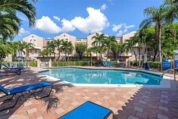 Outdoor swimming pool area with surrounding palm trees and a building in the background.