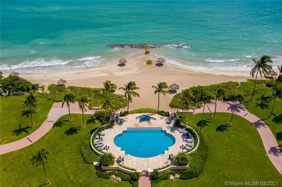 Aerial view of a beachfront property with a large swimming pool, surrounded by green lawns and palm trees, adjacent to a sandy beach and ocean.