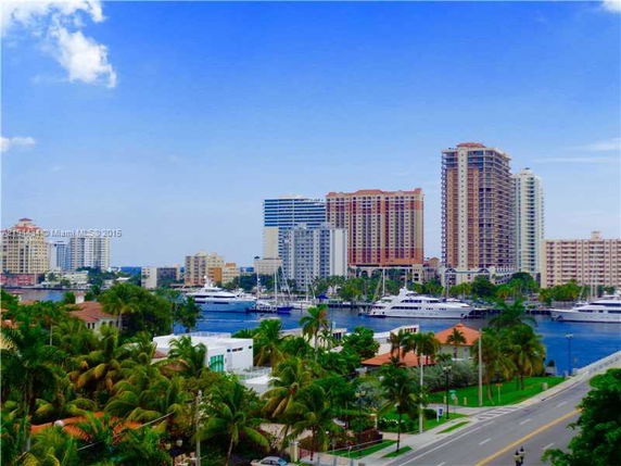 View of multiple high-rise buildings near a waterfront with yachts and palm trees in the foreground.