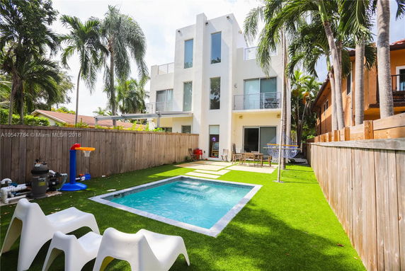 View of a modern three-story building with a backyard featuring a small swimming pool, chairs, and a fence.