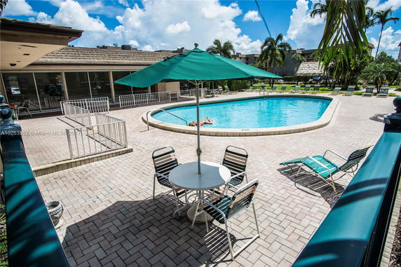 Outdoor pool area with a circular swimming pool, surrounded by lounge chairs, a green parasol, and patio furniture next to a building.