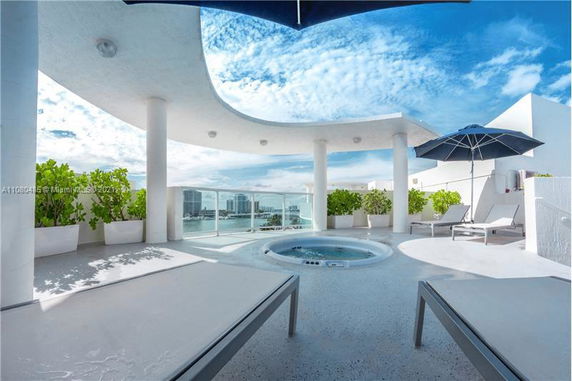 Rooftop area with a jacuzzi and lounge chairs, enclosed by glass railings, offering views of the cityscape.