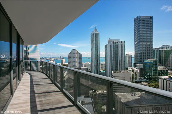 Panoramic view from a high-rise building balcony overlooking a cityscape with tall buildings and a distant body of water.