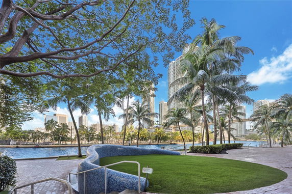 View of a cityscape with tall buildings and palm trees, seen from a park area with a water feature.