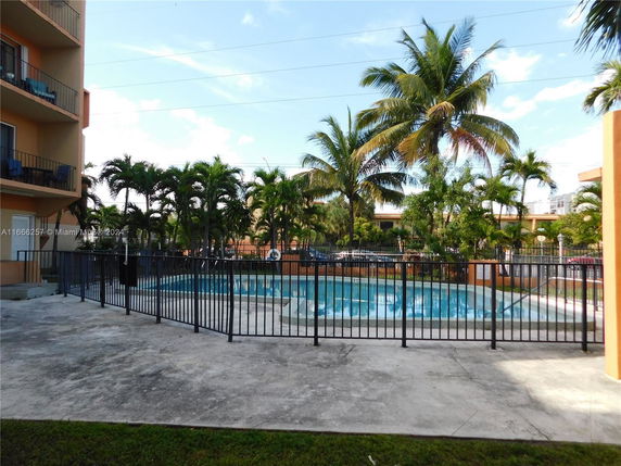 Outdoor swimming pool surrounded by a fence with nearby palm trees and building.