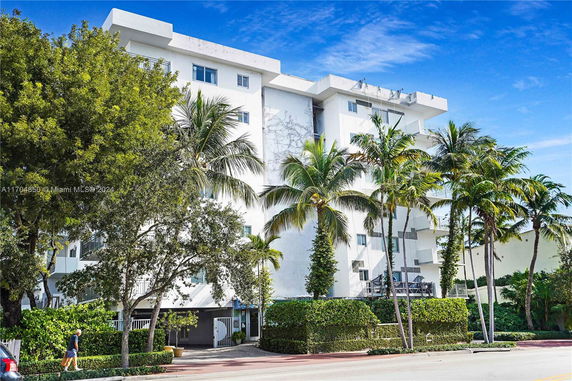 Front view of a multi-story white building with balconies and surrounded by palm trees.