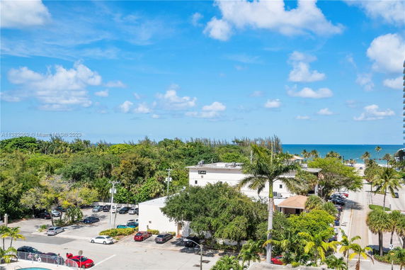 View of a small parking area, buildings, and a distant ocean with trees and palm trees.