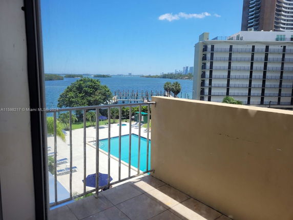 Balcony view overlooking a swimming pool with a waterfront and buildings in the background.