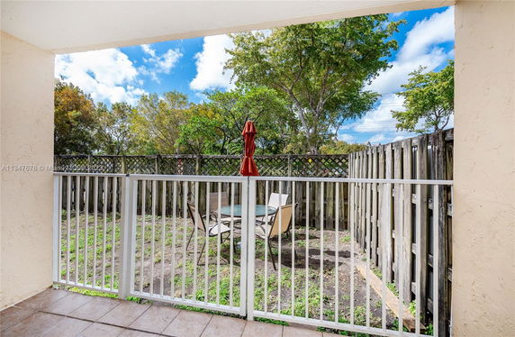 View from a patio showing a fenced backyard with outdoor furniture and trees beyond the fence.