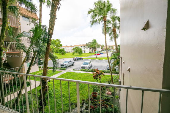 View from a balcony overlooking a parking area with palm trees and apartment buildings in the distance.