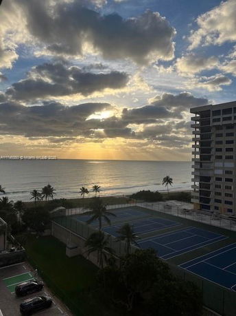 View of a coastal area with a high-rise building and tennis courts, overlooking the ocean at sunset.