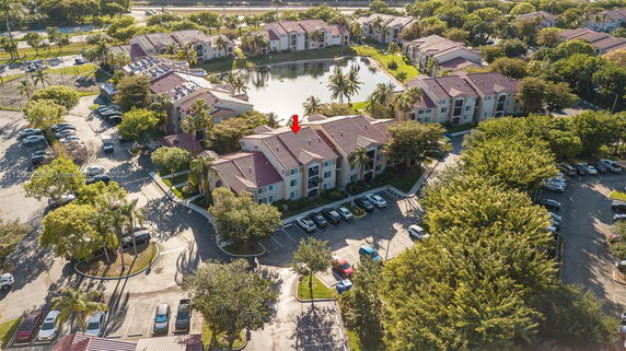 Aerial view of apartment complexes with parking areas, surrounded by trees and a pond.
