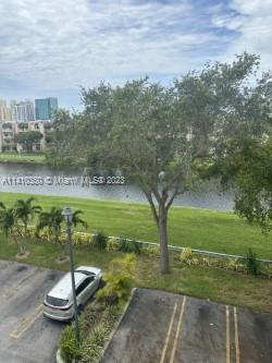 View of a parking area with a tree and greenery beside a water body, buildings visible in the background.