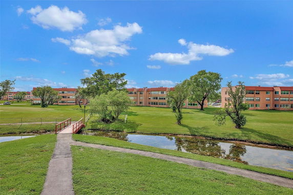 View of a multi-story residential building seen from across a lawn with trees and a small bridge over water.