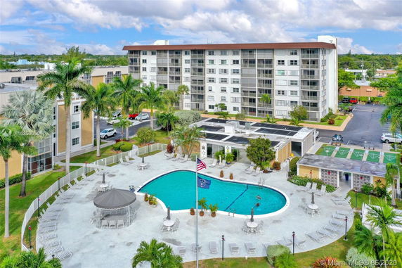 Outdoor swimming pool area with a circular arrangement of lounge chairs and umbrellas in front of a multi-story residential building.