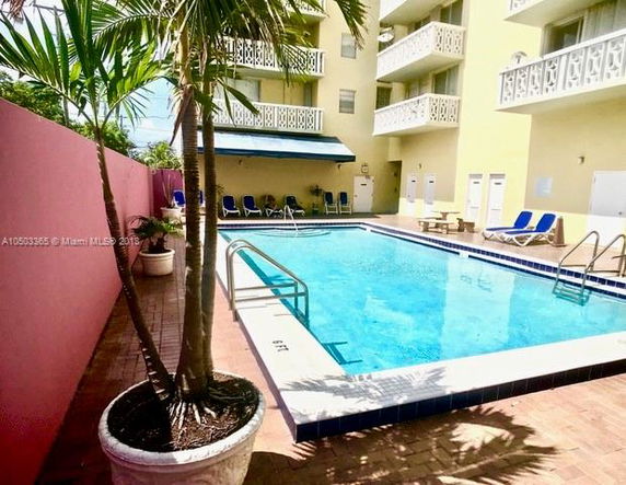 Pool area adjacent to a multi-story building with balconies and a pink wall.
