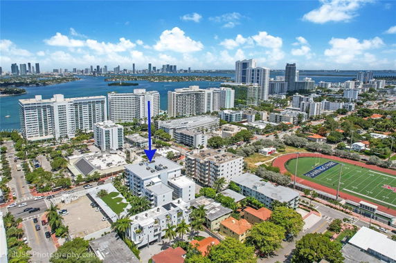 Aerial view of a coastal cityscape with multiple residential buildings, a sports field, and a body of water in the background.