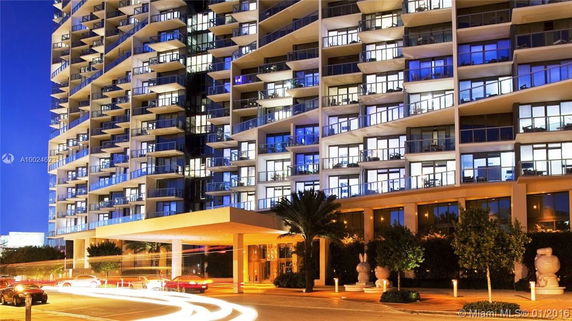 Front view of a multi-story apartment building with balconies and large windows, illuminated at night.