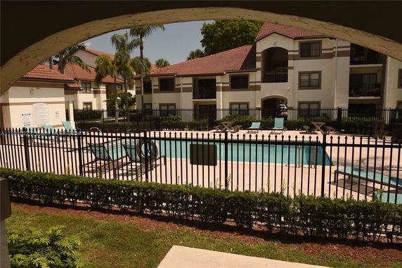 Outdoor view of a swimming pool with lounge chairs, surrounded by a metal fence, and apartment buildings in the background.