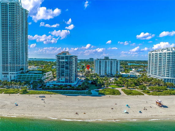 Aerial view of beachfront buildings with surrounding greenery and a sandy beach in the foreground.