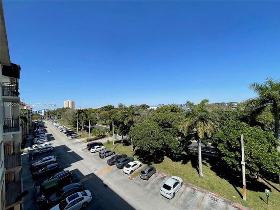 Outdoor parking area with multiple cars parked along a road lined with trees and buildings.