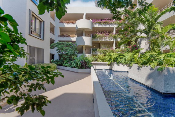 Outdoor view of a multi-story building with balconies adorned with plants, surrounded by greenery and a water feature.