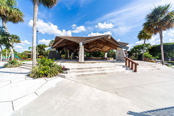 Open pavilion structure with wooden roof and stone pillars in a park setting.