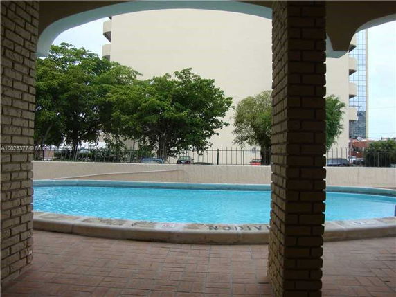 View of a swimming pool with brick pillars and a fence, surrounded by trees and buildings.
