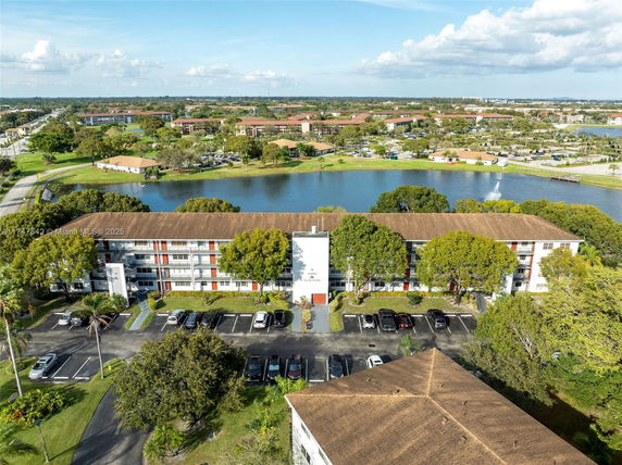 Aerial view showing a multi-story building with a parking area, surrounded by trees and a water body in the background.