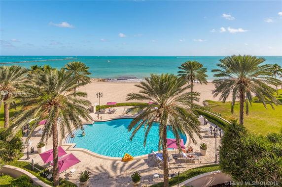 Panoramic view of a beach with a swimming pool, palm trees, and ocean in the background.