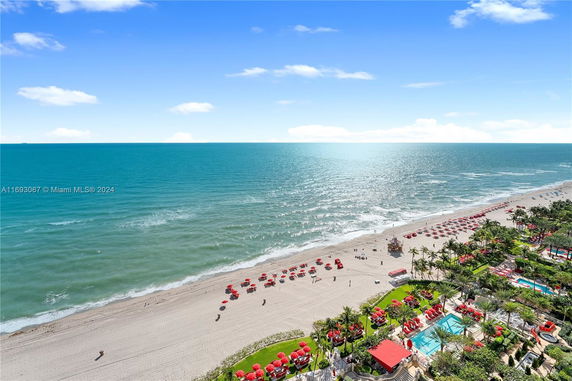Aerial view overlooking a beach with red umbrellas and sunbathers on the sandy shore, adjacent to a lush resort area.