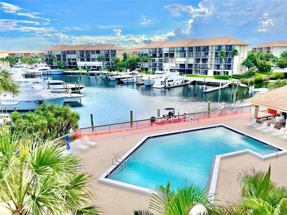 Panoramic view of a marina with several yachts and multi-story buildings in the background, seen from an elevated position with a swimming pool in the foreground.