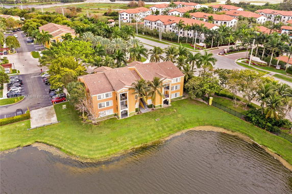 Aerial view of a multi-story residential building with brown roofs, surrounded by palm trees and adjacent to a body of water.