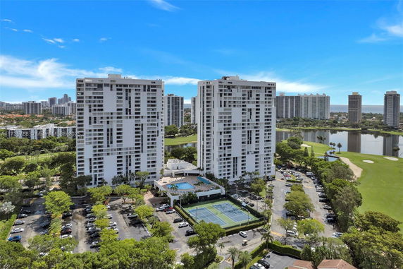 View of two high-rise residential buildings with a parking area and tennis court in the foreground.