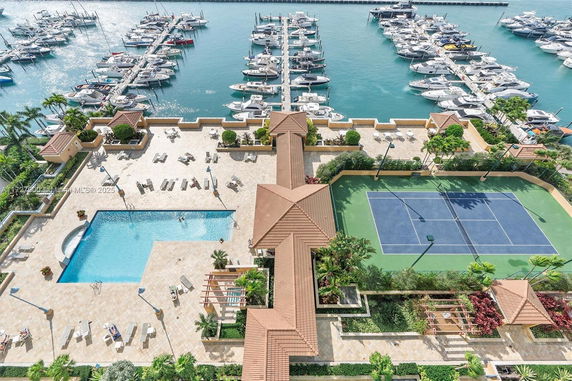 Panoramic view of outdoor area with swimming pool and tennis court next to a marina filled with boats.