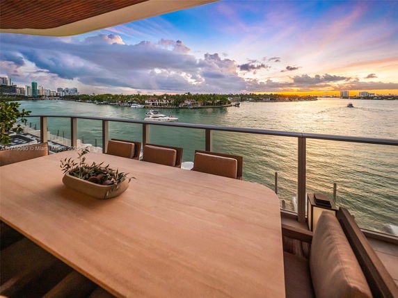 Outdoor panoramic view from a balcony overlooking a body of water with boats, and a cityscape in the distance during sunset.