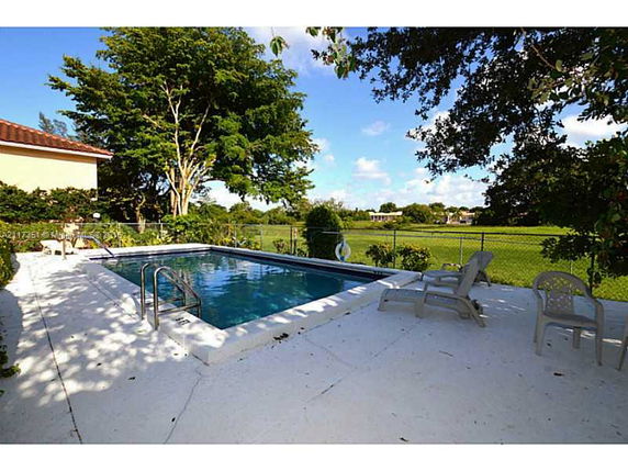 Backyard view of a swimming pool next to a house, with trees and open green space in the background.