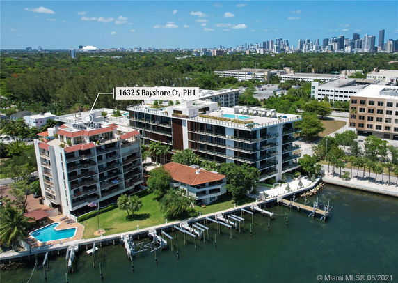 Aerial view of a waterfront condominium complex with docks and a swimming pool, set against a backdrop of a city skyline.