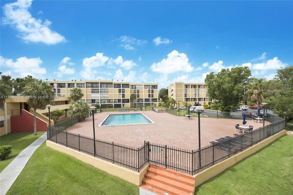 Outdoor view featuring a swimming pool surrounded by a paved area and fenced enclosure, with multi-story residential buildings in the background.