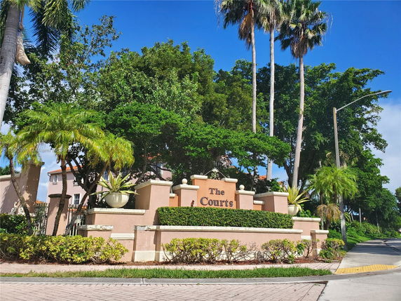 Entrance view of multi-unit residential complex named 'The Courts' with tropical landscaping and trees.