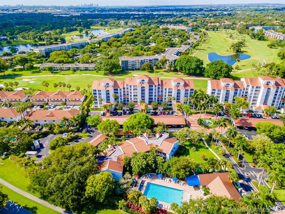 Aerial view of a residential complex with multiple multi-story buildings, surrounded by trees, a pool, and expansive green areas.